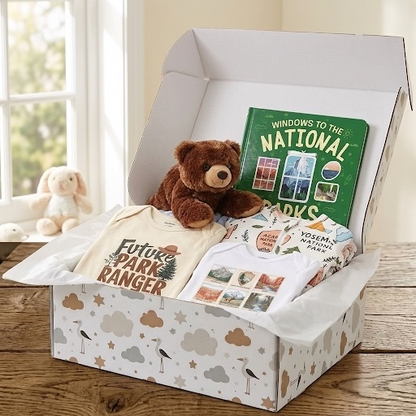 A curated gift box set displayed on a wooden table in a bright room. The open white patterned box contains a cream t-shirt reading 'Future Park Ranger,' a brown teddy bear, a green 'Windows to the National Parks' book, and a white t-shirt with Yosemite-themed graphics.