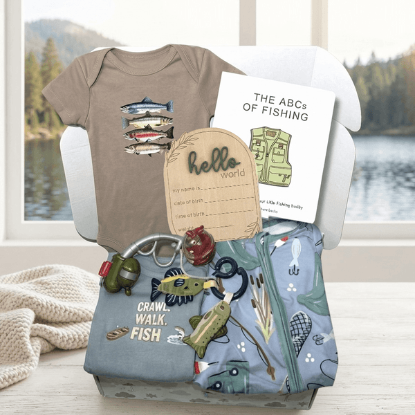 A baby gift basket arranged in a white chair, featuring a taupe short-sleeve baby bodysuit with a fish graphic, a white 'The ABCs of Fishing' book, a wooden 'hello world' card, a light blue zippered pouch with fishing-themed prints, and a small plush fish toy, set against a soft-focus lake and forest background.