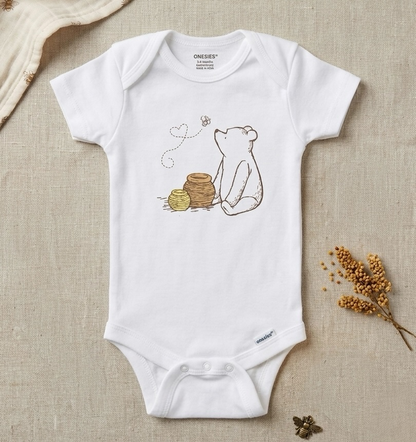 A white short-sleeve baby bodysuit laid flat on a beige linen background, featuring a printed illustration of a bear sitting beside a honey pot with a bee and heart detail. The brand name 'ONEEES' is visible inside the collar.