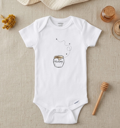 A white short-sleeve baby bodysuit laid flat on a beige linen surface, featuring a printed honey jar graphic with the word 'Honey' and a small bee. The bodysuit is styled with dried flowers, honey jars, and a wooden honey dipper.