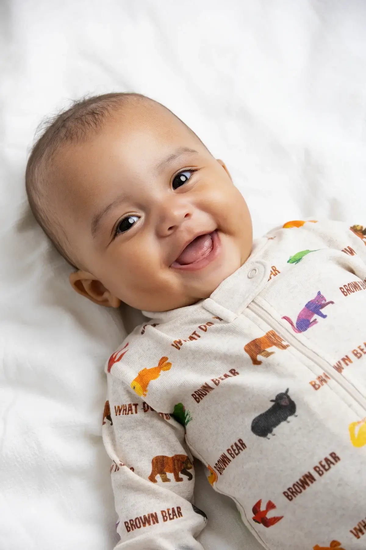 A smiling baby lying on white bedding, wearing a light beige long-sleeve footless onesie printed with colorful animal illustrations and the words 'BROWN BEAR' and 'WHAT A TIME PIG'.