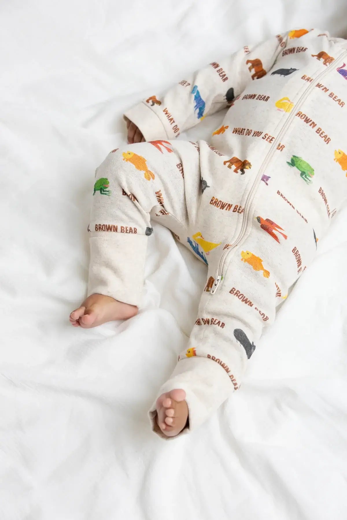 A baby lying on white bedding wearing a cream-colored zip-up sleeper with colorful animal prints and 'BROWN BEAR' text.