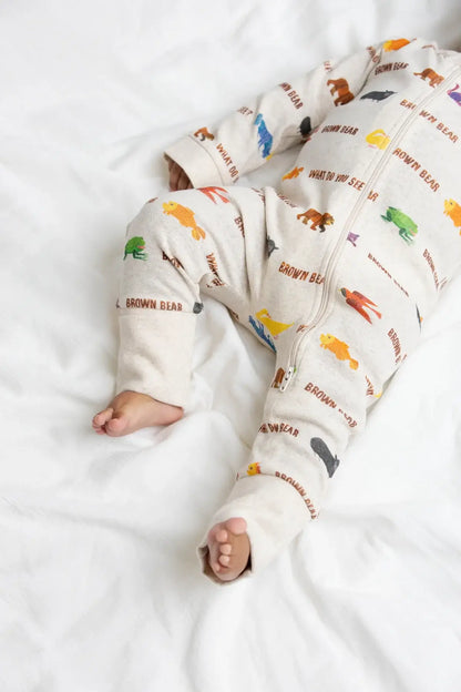 A baby lying on white bedding wearing a cream-colored zip-up sleeper with colorful animal prints and 'BROWN BEAR' text.