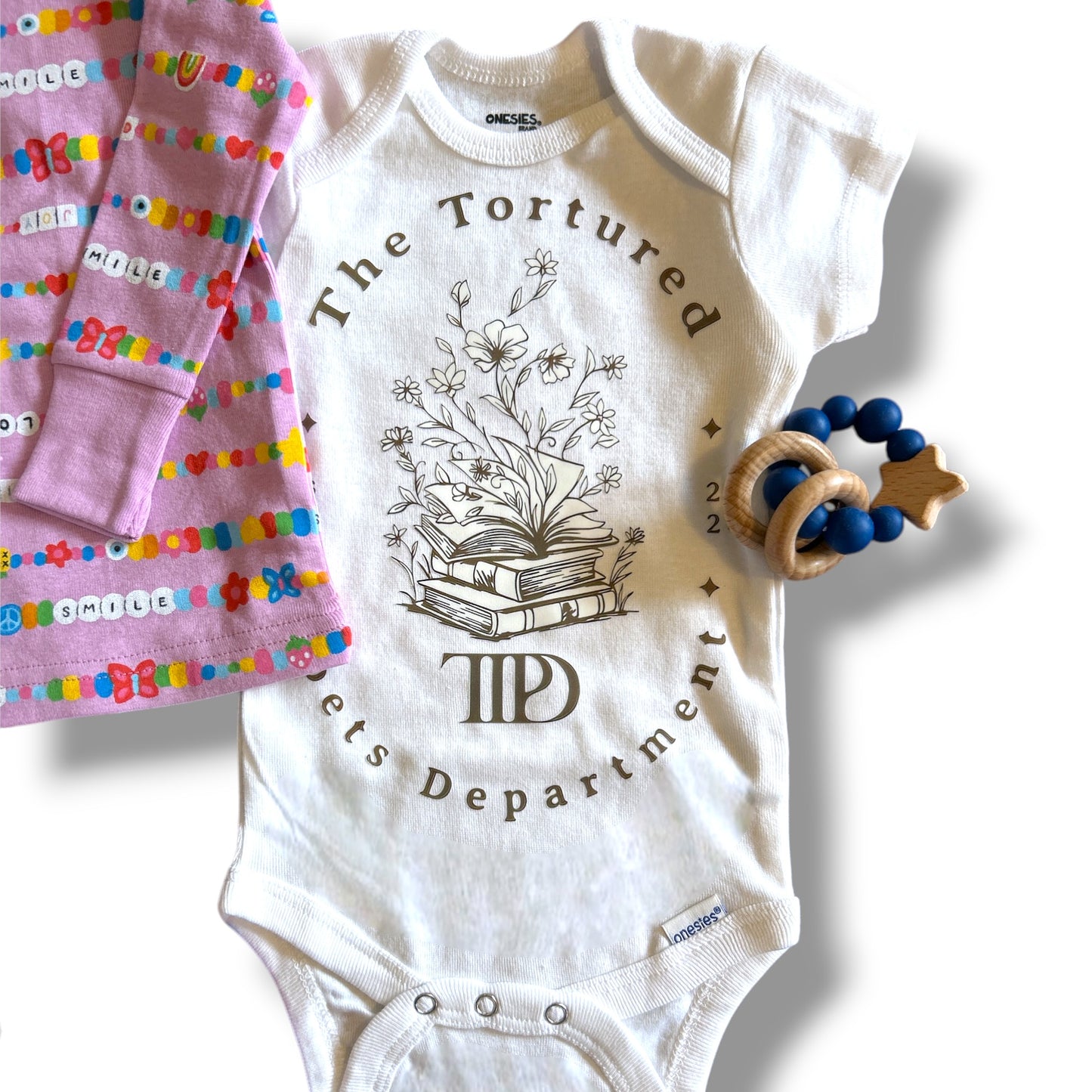 White baby onesie with 'The Tortured Poet's Department' design, pink baby blanket with colorful letters, and wooden teething ring on a white background.
