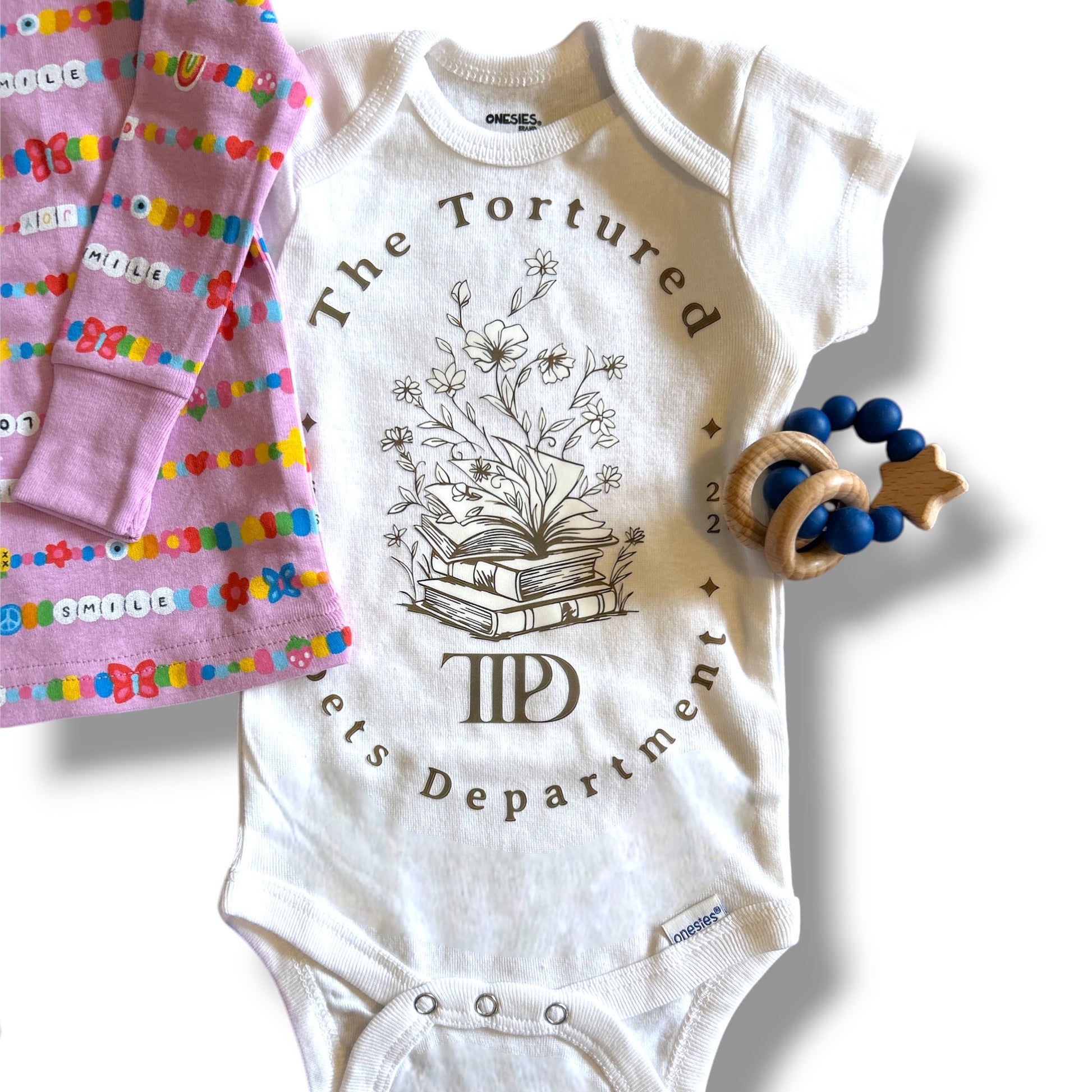 White baby onesie with 'The Tortured Poet's Department' design, pink baby blanket with colorful letters, and wooden teething ring on a white background.