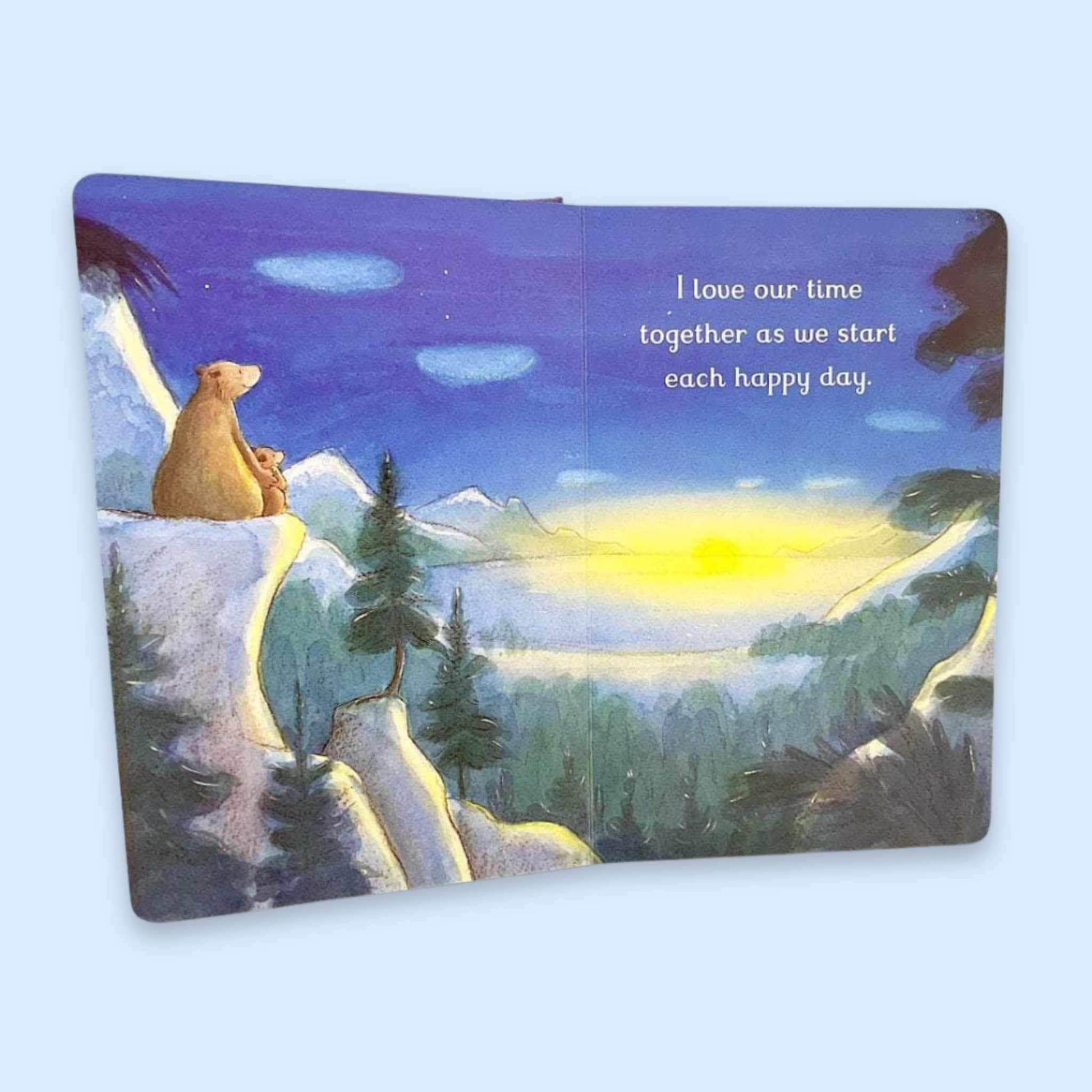 An open children's book showing a painted winter landscape with a bear sitting on a snowy cliff, mountains, trees, and a glowing sun. White text on the right page reads, 'I love our time together as we start each happy day.