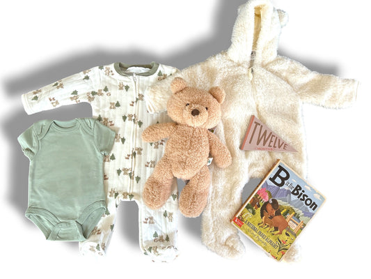 Baby clothing and toys including a onesie, teddy bear, blanket, and book on a white background