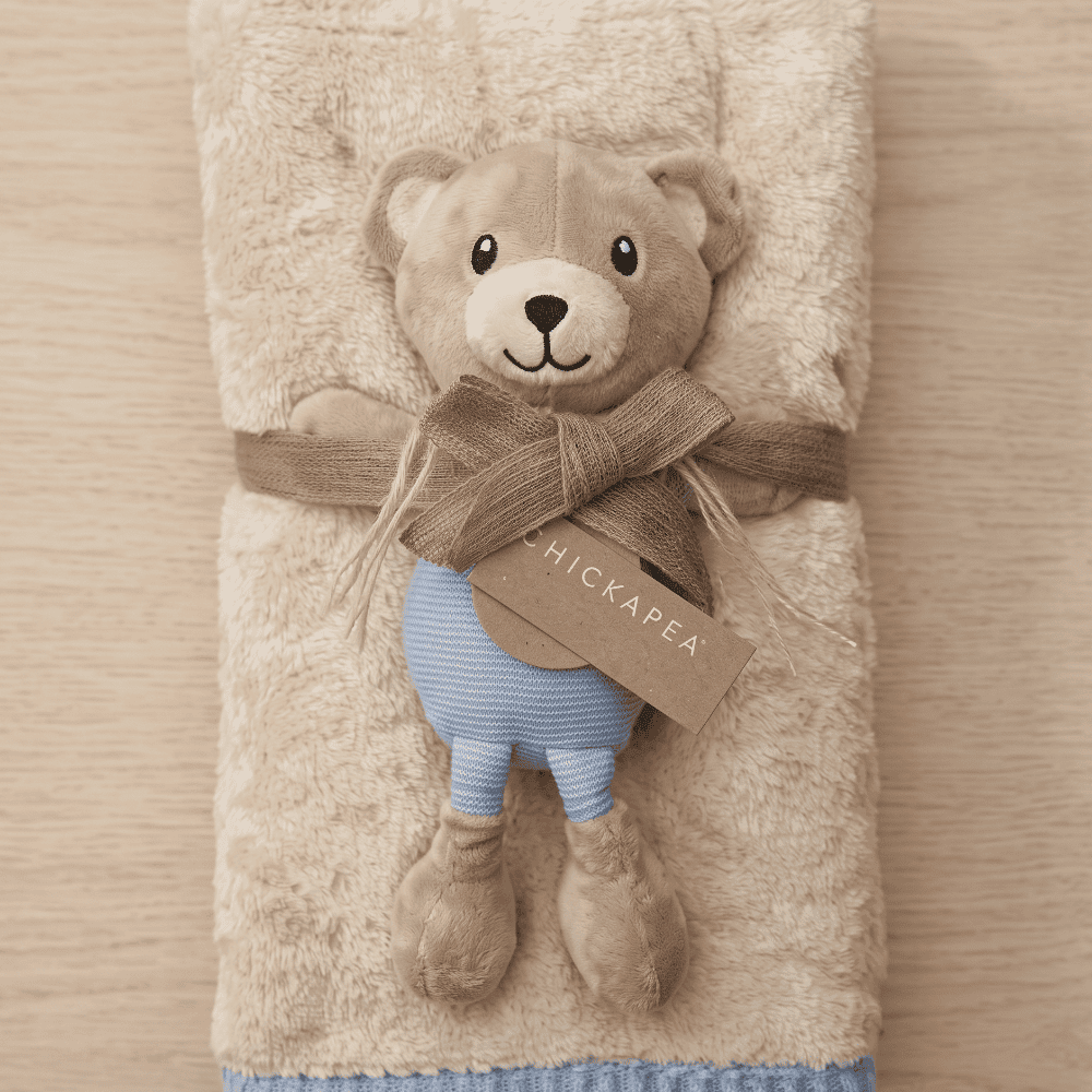 Teddy bear plush toy with a scarf and blue pants, wrapped in a beige blanket on a wooden surface.