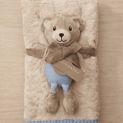 Teddy bear plush toy with a scarf and blue pants, wrapped in a beige blanket on a wooden surface.