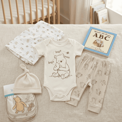 Baby clothing set with Winnie the Pooh design on a couch next to a crib and books.