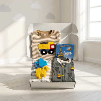 A baby gift set in an open white box, featuring a beige striped long-sleeve shirt with a yellow construction truck applique, a gray long-sleeve romper with yellow construction equipment prints, a blue book titled 'Goodnight Construction Site', and two yellow and blue plush toys, set in a bright room with window light.