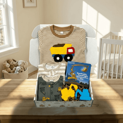 A baby gift box displayed on a wooden table in a softly lit nursery. The box contains a striped baby shirt with a yellow construction truck applique, a gray onesie, a blue children's book titled 'Goodnight, Construction Site,' and yellow and blue toy pieces.