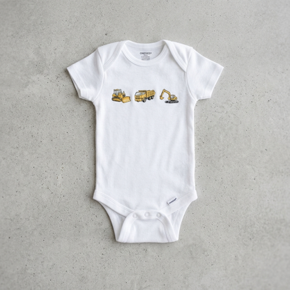 A white short-sleeve baby bodysuit laid flat on a textured gray concrete surface. The bodysuit features a graphic print of three construction vehicles in yellow and gray on the chest: a bulldozer, a dump truck, and an excavator.