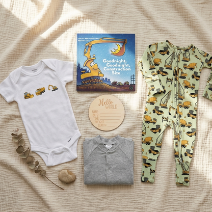 A baby gift set arranged on a textured cream blanket with soft sunlight. The set includes a white short-sleeve bodysuit with construction vehicle prints, a light green one-piece with a yellow construction vehicle print, a gray folded long-sleeve top, a children's book titled 'Goodnight, Goodnight, Construction Site,' a round wooden name card, and a small eucalyptus sprig.