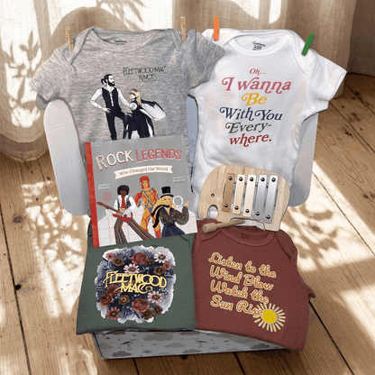 A curated baby gift set arranged on a wooden floor with sunlight and shadow overlays. The set includes a gray and white crewneck baby bodysuit set, a 'Rock Legends' book, a wooden toy, and two crewneck shirts with Fleetwood Mac graphics.