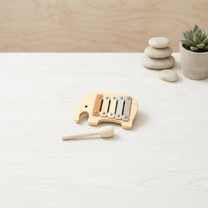 A small wooden elephant-shaped xylophone toy with metal bars and a matching wooden mallet, placed on a light wood tabletop with stacked stones and a small potted succulent in the background.