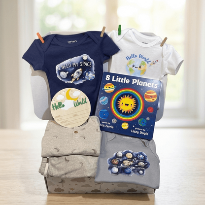 A baby gift set displayed on a light wood surface in front of a softly blurred window. The set includes a navy blue baby bodysuit with a space-themed graphic, a white baby bodysuit with a cartoon planet graphic, a folded light gray patterned blanket, a round cream sticker, and a children's book titled '8 Little Planets' by Laura Ferris.
