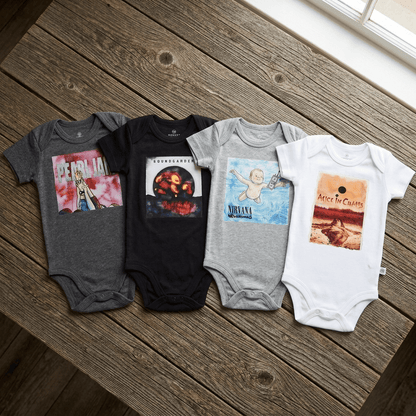 Four baby bodysuits laid out on a rustic wooden floor, each printed with a different Nirvana graphic design. The bodysuits are in gray, black, light gray, and white, featuring the band's 'In Bloom', 'Bloomer Bringer', 'Lifefist', and 'Peace Sells' album art.