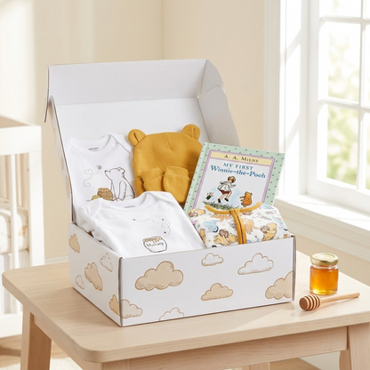 A curated baby gift box set displayed on a light wood table in a bright nursery. The set includes a white baby bodysuit with a cartoon bear, a yellow plush toy, a children's book titled 'Winnie-the-Pooh', a patterned fabric item, and a small jar of honey with a wooden dipper.