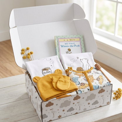 A baby gift box set displayed on a light wooden table near a window. The open white box contains a yellow knit bonnet, a white cloth with a bee illustration labeled 'Maisy', a Winnie-the-Pooh book, and a baby honey pot holder.