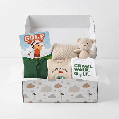 A baby gift box set containing a green zip-up baby outfit, a beige knit hat, a white onesie with green text, a teddy bear, and a 'Golf Baby' book, all arranged inside a patterned gift box on a white background.