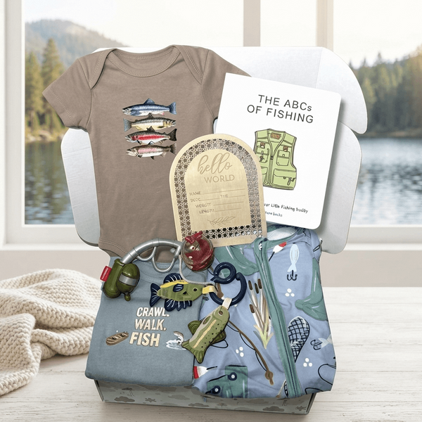A baby gift set displayed in a white box on a wooden table, featuring a taupe short-sleeve baby bodysuit with a fish graphic, a white card titled 'THE ABCS OF FISHING', a light blue zippered pouch with fishing-themed prints and a keychain, and a small woven tag. A window behind shows a lake and trees, and a cream knit blanket rests to the left.