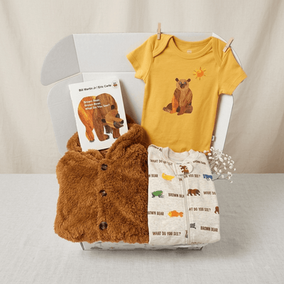 A baby gift set arranged in a clear box against a neutral fabric backdrop. The set includes a mustard yellow short-sleeve baby bodysuit with a brown bear graphic, a plush brown bear-themed card or wearable piece, a cream baby shirt with colorful animal illustrations, and a small book.