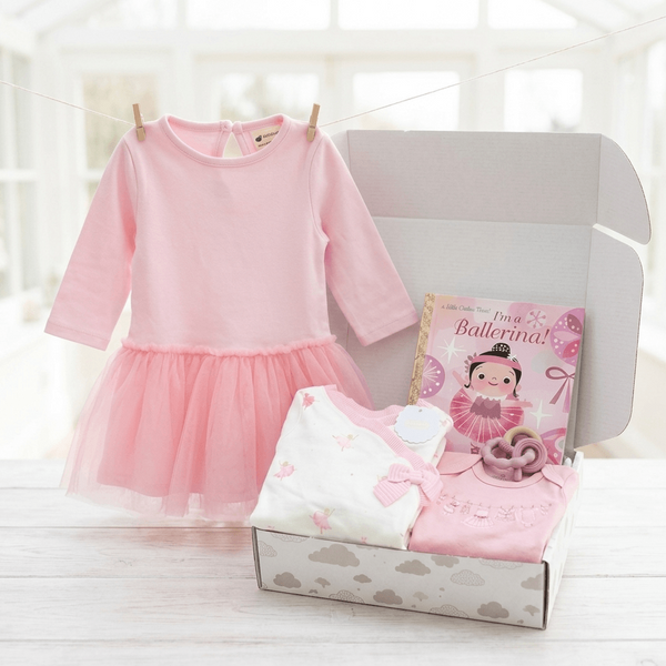A baby ballerina gift set displayed on a light wood table, featuring a pink long-sleeve tutu dress hanging on a string, a white and pink folded bodysuit, a pink pacifier, and a 'My First Ballerina!' book inside an open gray box.