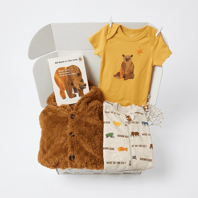 A baby gift set arranged in a white box on a light gray background, including a mustard yellow short-sleeve bodysuit with a bear graphic, a brown plush cardigan, a cream pajama set with bear prints, and a small book titled 'Bill Nye at the Camp Fire'.