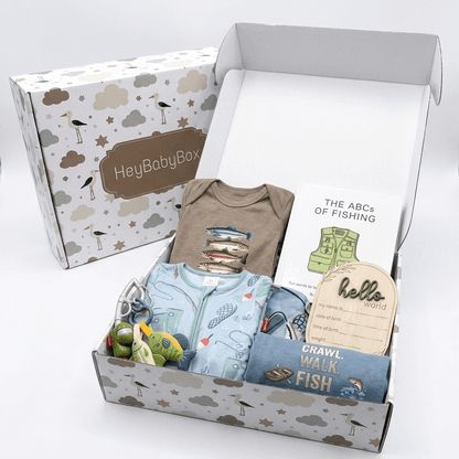 An open HeyBabyBox gift box filled with baby items, including a light blue onesie, a brown bodysuit, a fishing-themed card, a wooden name tag, and small accessories, with the branded patterned box behind it on a white background.