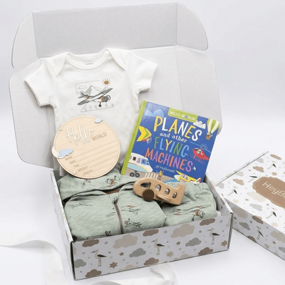 A baby gift set in a white cardboard box, featuring a white short-sleeve baby bodysuit with an airplane graphic, a light green patterned zip-up romper, a blue book titled 'Planes and Other Flying Machines', a wooden toy, and a round wooden tag. A matching branded gift box sits beside the open box on a white background.