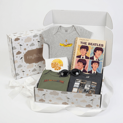 A baby gift box set containing a gray baby bodysuit with a yellow submarine graphic, a green t-shirt, a dark gray t-shirt with band posters, a Beatles book, a white t-shirt, and sunglasses, all arranged inside an open patterned box next to its branded box.