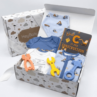 A baby gift set in a printed cardboard box, featuring a blue and white onesie, a light blue bib, a dark gray construction-themed book, and toy tools in orange, yellow, and blue, with a HeyBaby box in the background.