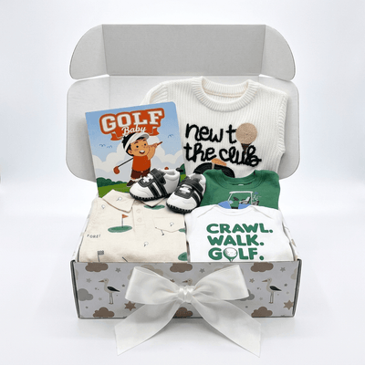 A baby gift set in an open gray box with a white ribbon bow, featuring a white long-sleeve shirt that says 'newt the club', a green short-sleeve shirt, folded cream and white garments with golf-themed prints, a pair of black-and-white booties, and a 'GOLF Baby' card.