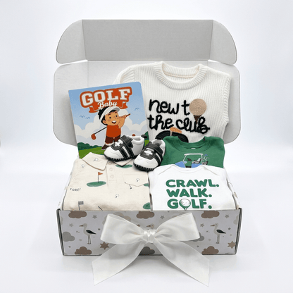 A baby gift set in an open gray box with a white ribbon bow, featuring a white long-sleeve shirt that says 'newt the club', a green short-sleeve shirt, folded cream and white garments with golf-themed prints, a pair of black-and-white booties, and a 'GOLF Baby' card.