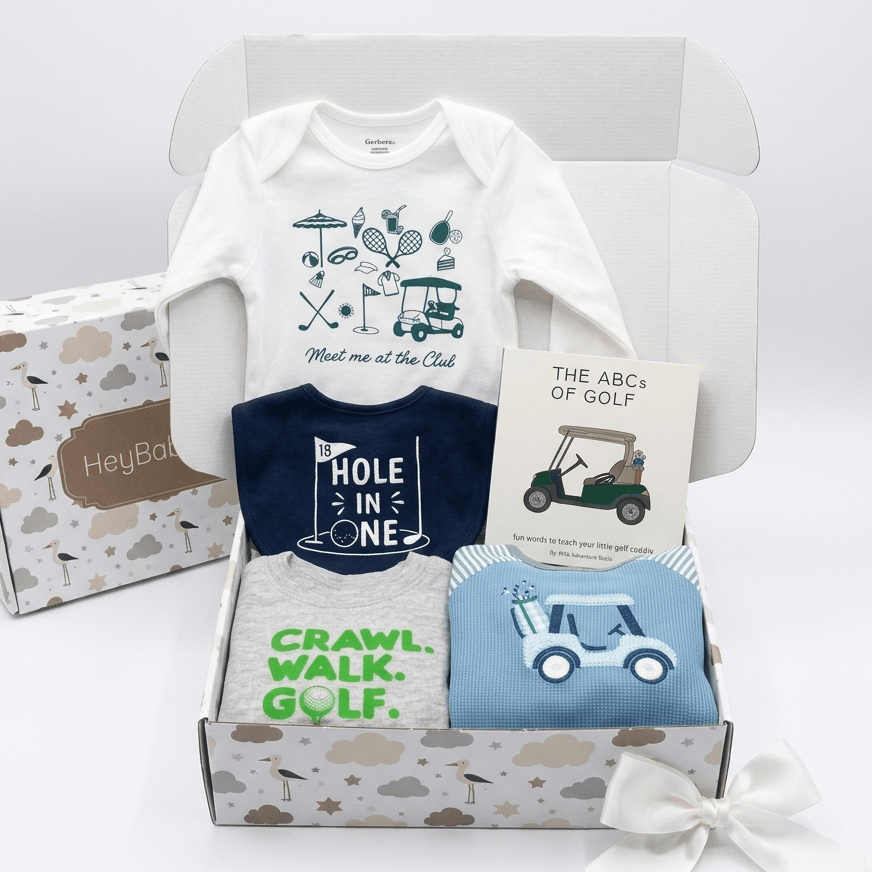 A baby gift set in a white box featuring a white long-sleeve bodysuit with a golf-themed graphic, a navy blue shirt that says 'HOLE IN ONE', a light blue folded garment with a golf cart graphic, a card reading 'THE ABCS OF GOLF', and a patterned gift box with a white bow.