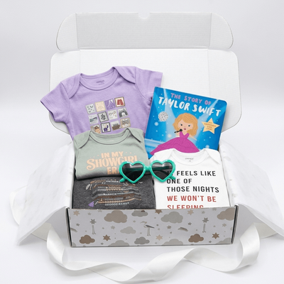 A curated baby gift box set displayed inside an open white box with tissue paper and a white ribbon. The set includes a lavender short-sleeve baby bodysuit, a sage green bodysuit with 'In My Shower I Won' text, a white bodysuit with 'Feels Like One of Those Nights We Won't Be Sleeping' text, a dark gray t-shirt, a purple heart-shaped sunglasses, and a children's book titled 'The Story of Taylor Swift'.