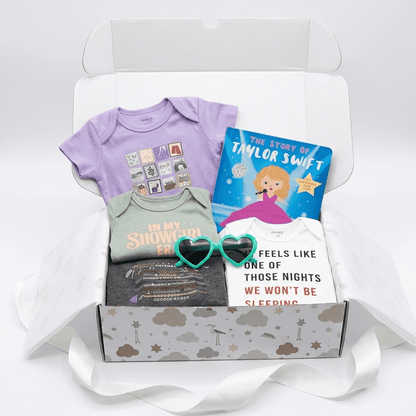 A curated baby gift box set displayed inside an open white box with tissue paper and a white ribbon. The set includes a lavender short-sleeve baby bodysuit, a sage green bodysuit with 'In My Shower I Won' text, a white bodysuit with 'Feels Like One of Those Nights We Won't Be Sleeping' text, a dark gray t-shirt, a purple heart-shaped sunglasses, and a children's book titled 'The Story of Taylor Swift'.
