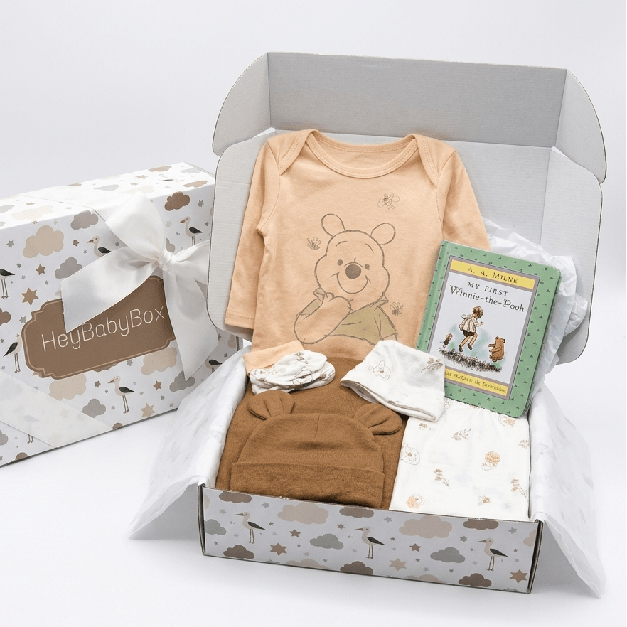 A baby gift set in an open white box with cloud and star print, including a peach long-sleeve bodysuit with Winnie the Pooh graphic, a brown top, folded white and tan textiles, a green 'My First Winnie the Pooh' book, and a wrapped 'HeyBabyBox' gift box beside it on a white background.