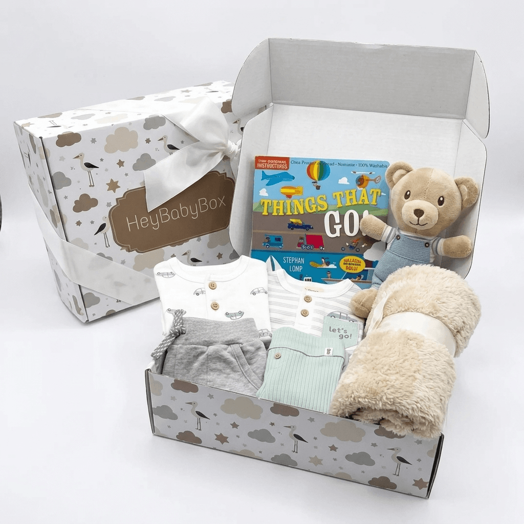 A baby gift set in a patterned box with a plush teddy bear, a book titled 'Things That Go', folded baby clothing, and a soft cream blanket. A branded outer box with a white ribbon sits behind the open gift box on a clean white background.