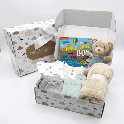 A baby gift set in a patterned box with a plush teddy bear, a book titled 'Things That Go', folded baby clothing, and a soft cream blanket. A branded outer box with a white ribbon sits behind the open gift box on a clean white background.