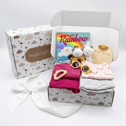 A baby gift box set from HeyBabyBox on a white background. The open box contains a pink long-sleeve romper, a light pink onesie, a gray cloth, a yellow ring toy, a plush bunny, a book titled 'The Sun Rainbow', a wooden 'Hello World' card, and a white satin bow.