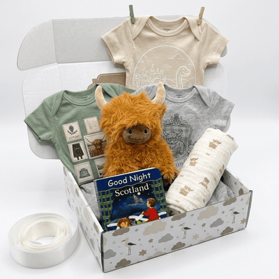 A baby gift box set containing a beige baby bodysuit, a green graphic t-shirt, a gray graphic t-shirt, a fluffy orange Highland cow plush toy, a white muslin blanket, a book titled 'Good Night Scotland', and a roll of white ribbon, all arranged in a patterned gift box on a white background.