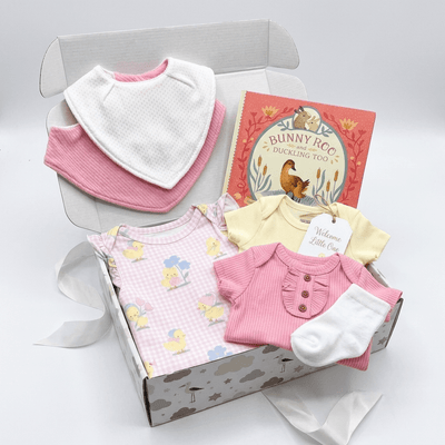 A baby gift set arranged inside an open gray box on a white background. The set includes a pink and white gingham baby top with bunny prints, a pink ribbed bodysuit, a white ribbed bib, a pale yellow short-sleeve bodysuit, a white sock, and a children's book titled 'Bunny Recess: Bucking Up!'.