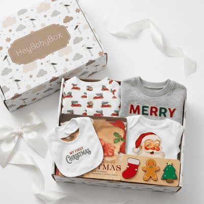 A Christmas-themed baby gift box set from HeyBabyBox, featuring a white patterned box lid, a white ribbon bow, and an open box containing baby clothing and accessories including a red truck print bodysuit, a gray 'MERRY' long-sleeve shirt, a white 'My First Christmas' bib, a Santa Claus bodysuit, and holiday-themed cards and stickers.