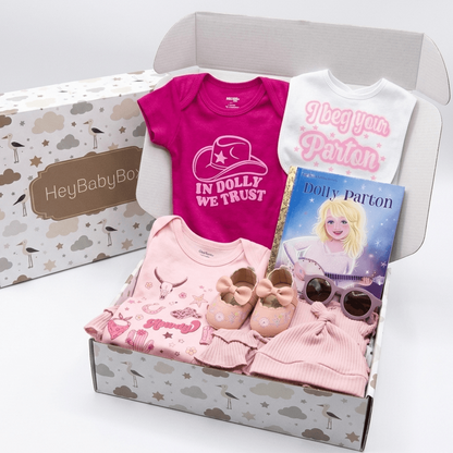 A baby gift box set from HeyBabyBox containing baby clothes and accessories. The open box contains a pink baby t-shirt with a cowboy hat graphic and the text 'IN DOLLY WE TRUST', a white baby bodysuit with 'I beg your pardon' in pink, a pink long-sleeve top, a pair of pink booties, and a 'Dolly Parton' book.
