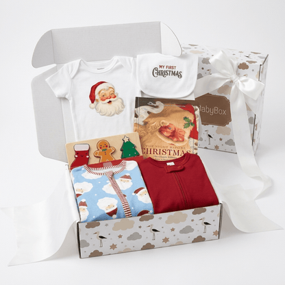 A Christmas-themed baby gift box set from BabyBox, shown open with a white onesie featuring Santa, a white bib reading 'My First Christmas,' a red zip-up sleeper, and a blue patterned outfit with Santa prints, arranged in a white gift box with a white ribbon bow.