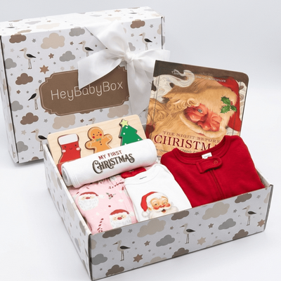 A Christmas-themed baby gift box set by HeyBabyBox, featuring a white ribbon tied to the box, a book titled 'The Naughty Bright Christmas', and folded baby clothes including a red top, a white shirt with Santa graphics, and a pink onesie, all arranged in an open patterned box.