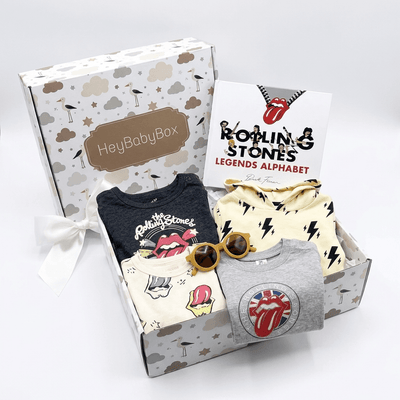 A baby clothing gift set from HeyBabyBox featuring a Rolling Stones themed alphabet set in a patterned box. The set includes a navy blue t-shirt, a cream long-sleeve shirt with black lightning bolts, a grey t-shirt with a red and white Rolling Stones tongue logo, a cream t-shirt with pink and yellow graphics, and a pair of yellow sunglasses, all arranged in front of the gift box and a brand card.