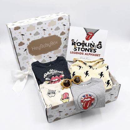 A baby clothing gift set from HeyBabyBox featuring a Rolling Stones themed alphabet set in a patterned box. The set includes a navy blue t-shirt, a cream long-sleeve shirt with black lightning bolts, a grey t-shirt with a red and white Rolling Stones tongue logo, a cream t-shirt with pink and yellow graphics, and a pair of yellow sunglasses, all arranged in front of the gift box and a brand card.